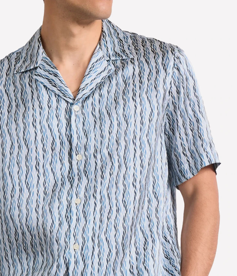 A close-up of a man wearing a Frescobol Carioca Roberto Short Sleeve Silk Shirt in Denim Blue, short-sleeve button-up shirt with a vertical wavy abstract pattern in light blue, white, and black.