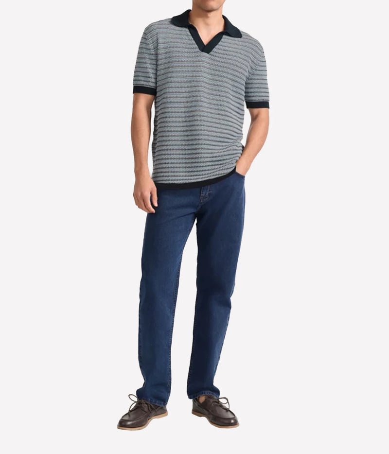 A man wearing a Frescobol Carioca Rino Short Sleeve Cotton Polo in Navy, dark blue jeans, and brown leather shoes, standing against a light grey background.