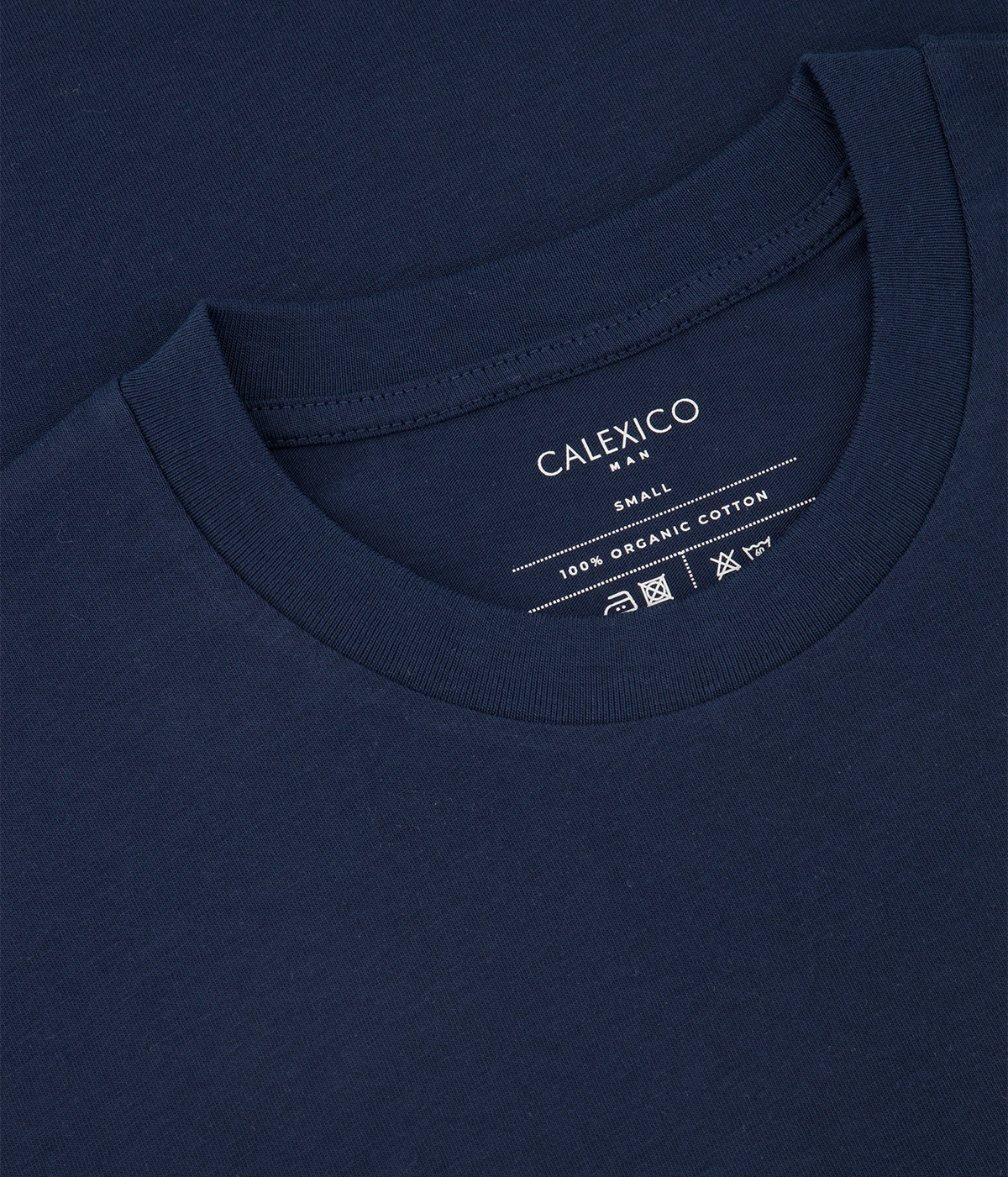 Close-up of a navy blue crewneck t-shirt showing the inner neck label with white text that reads 'CALEXICO MEN', 'SMALL', and '100% ORGANIC COTTON'.