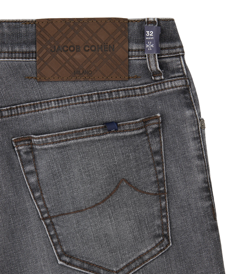 Close-up of the back waistband and pocket area of gray denim jeans, showing a brown jacquard brand patch that reads JACQUE COHEN MONO, a dark blue size tab marked 32, and contrast stitching.