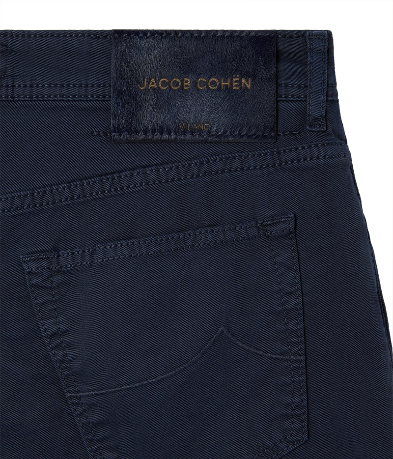 Close-up of the back waistband and pocket of dark navy blue jeans by Jacob Cohen, showing a branded leather patch and gold-tone embroidery on a white background.