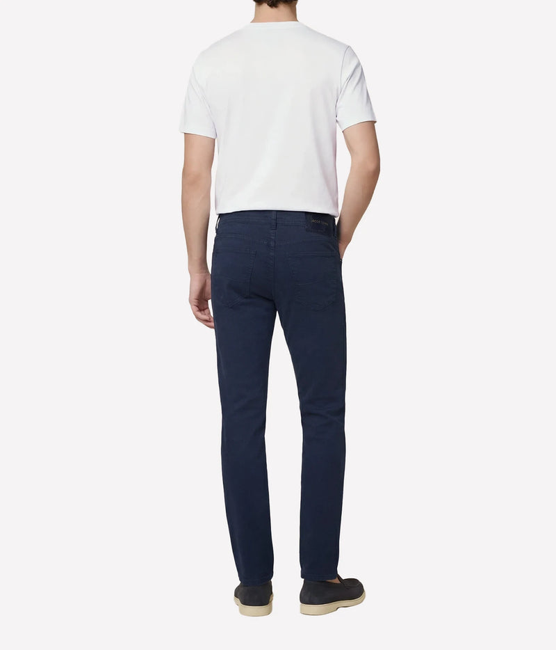 A man viewed from the back wearing a white short-sleeve t-shirt, navy blue slim-fit jeans, and dark slip-on shoes, set against a plain light gray studio background.