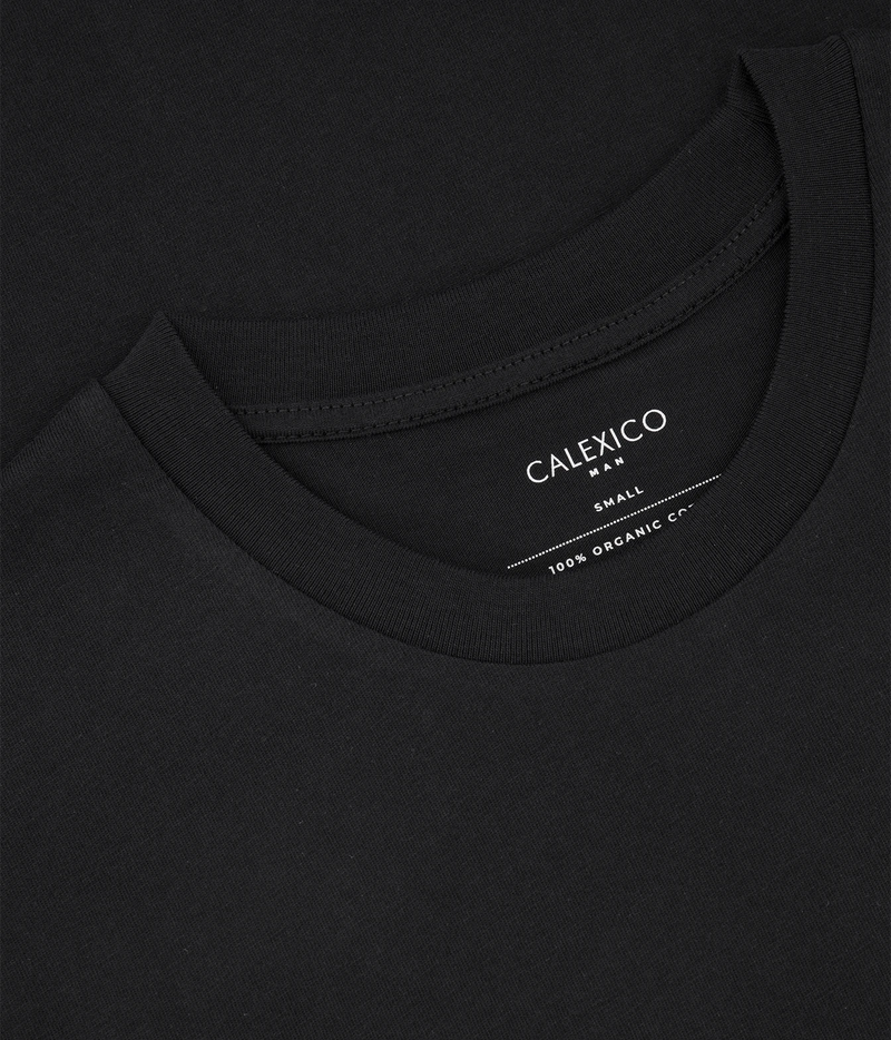Close-up of a black crewneck t-shirt showing the inner neck label with white text that reads 'CALEXICO SMALL 100% ORGANIC CO'.
