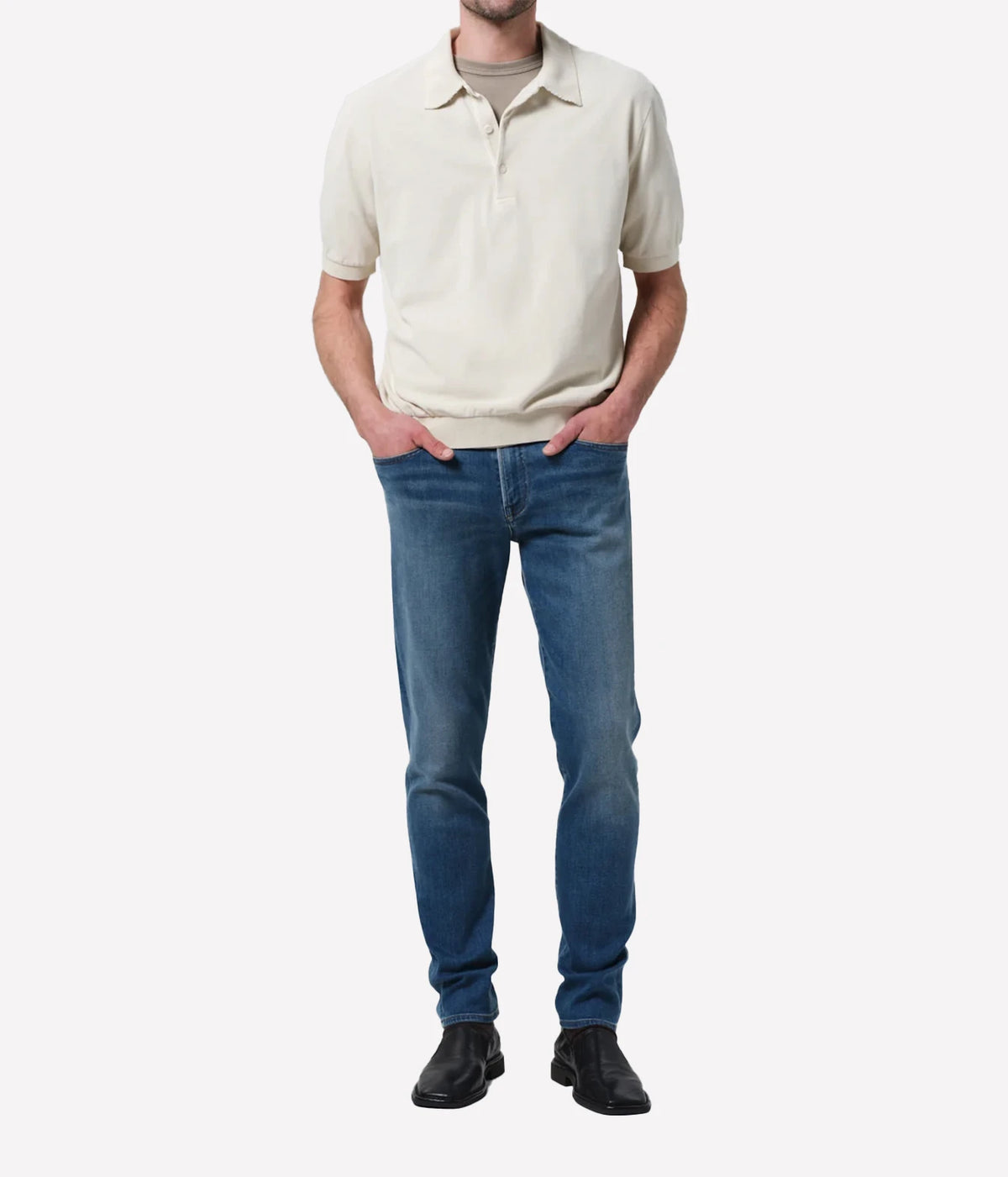 A man wearing a cream short-sleeve polo shirt, blue jeans, and black shoes, standing against a plain light grey background.