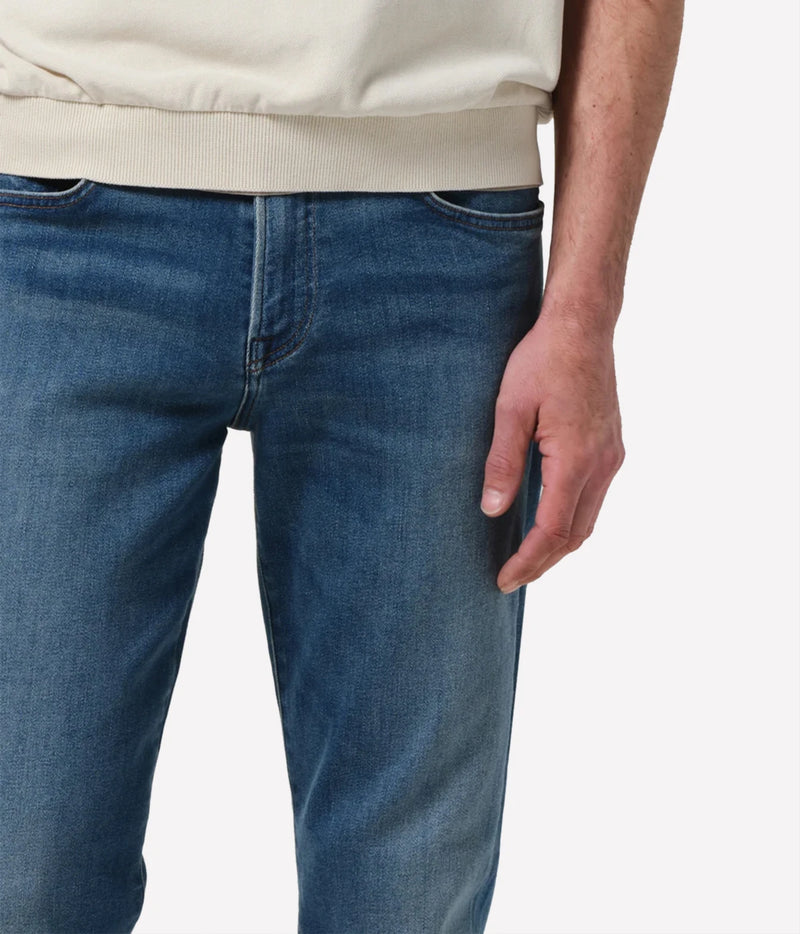 Close-up of a person wearing blue denim jeans and a cream sweatshirt, showing the front waistband, pocket, and hand against a plain light background.