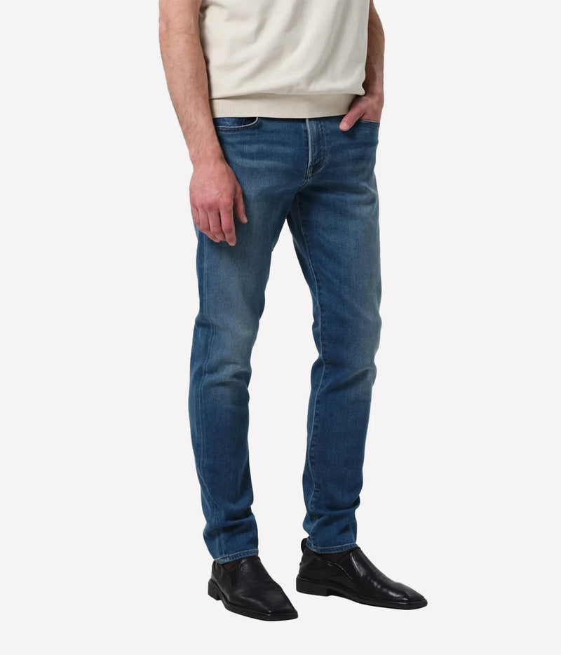 A person wearing slim-fit medium wash blue jeans, a cream sweatshirt, and black leather boots, shown from the waist down against a light grey studio background.