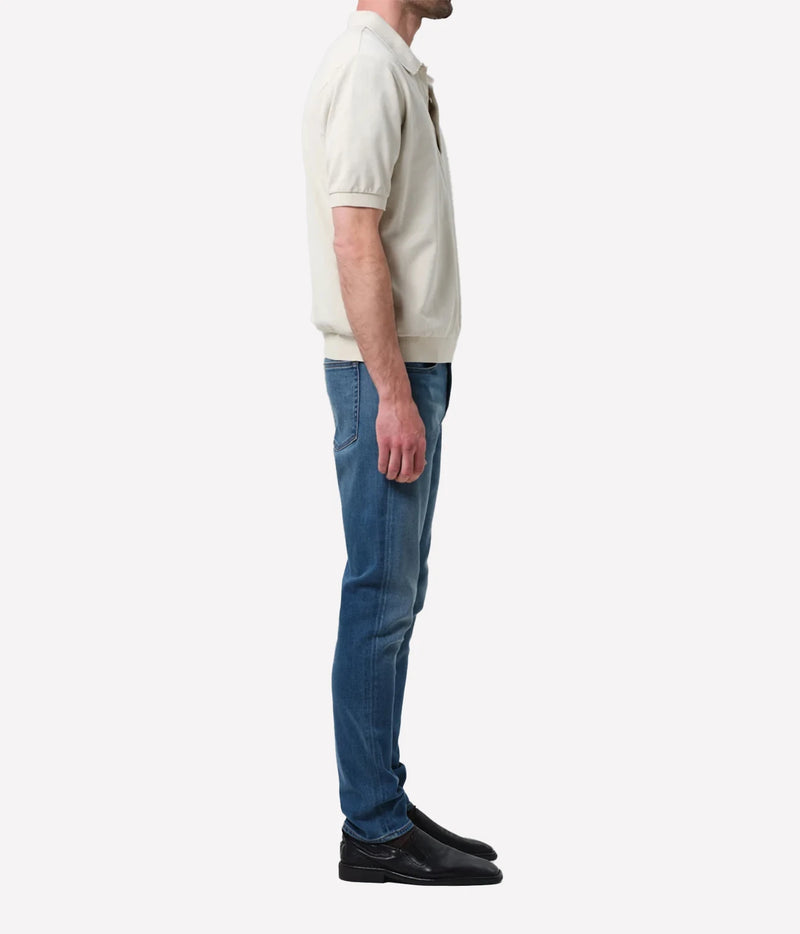 A man shown in profile wearing a cream short-sleeve polo shirt, blue jeans, and black shoes against a plain light grey studio background.