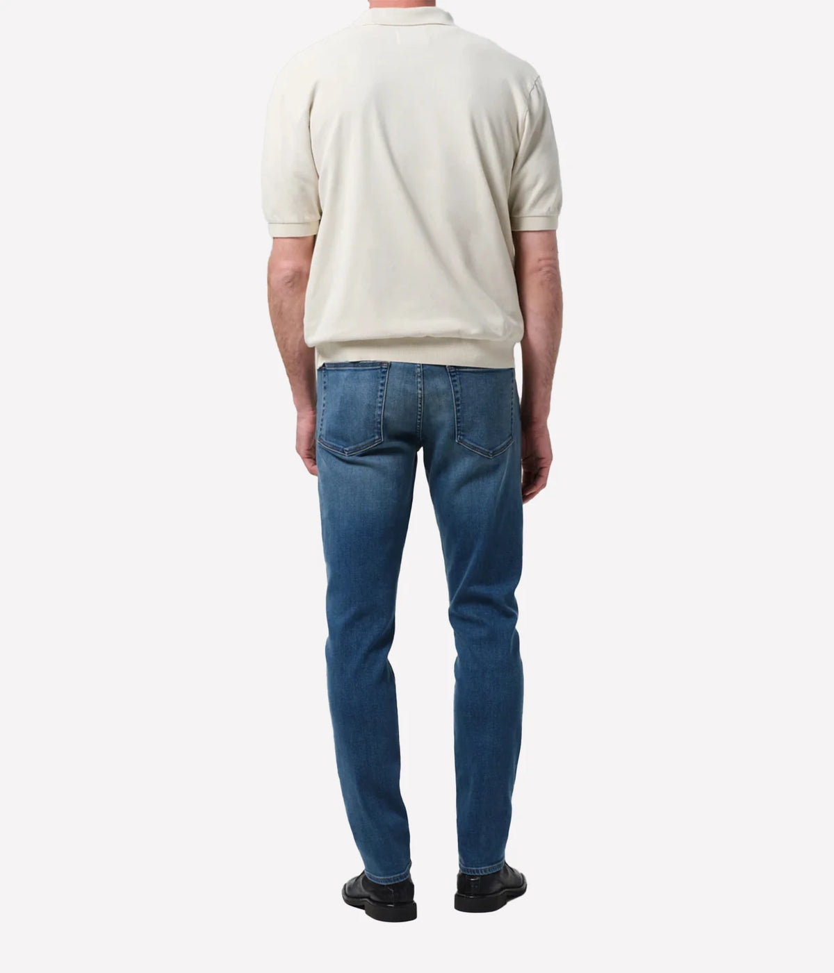 A man viewed from the back wearing a cream short-sleeve knit top and blue jeans, standing against a plain light grey studio background.