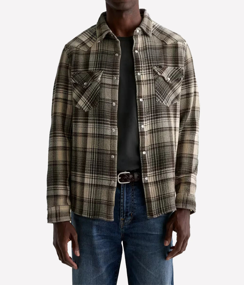 A man wearing a brown and beige plaid flannel shirt over a black t-shirt and blue jeans, shown from the neck down against a light gray background.