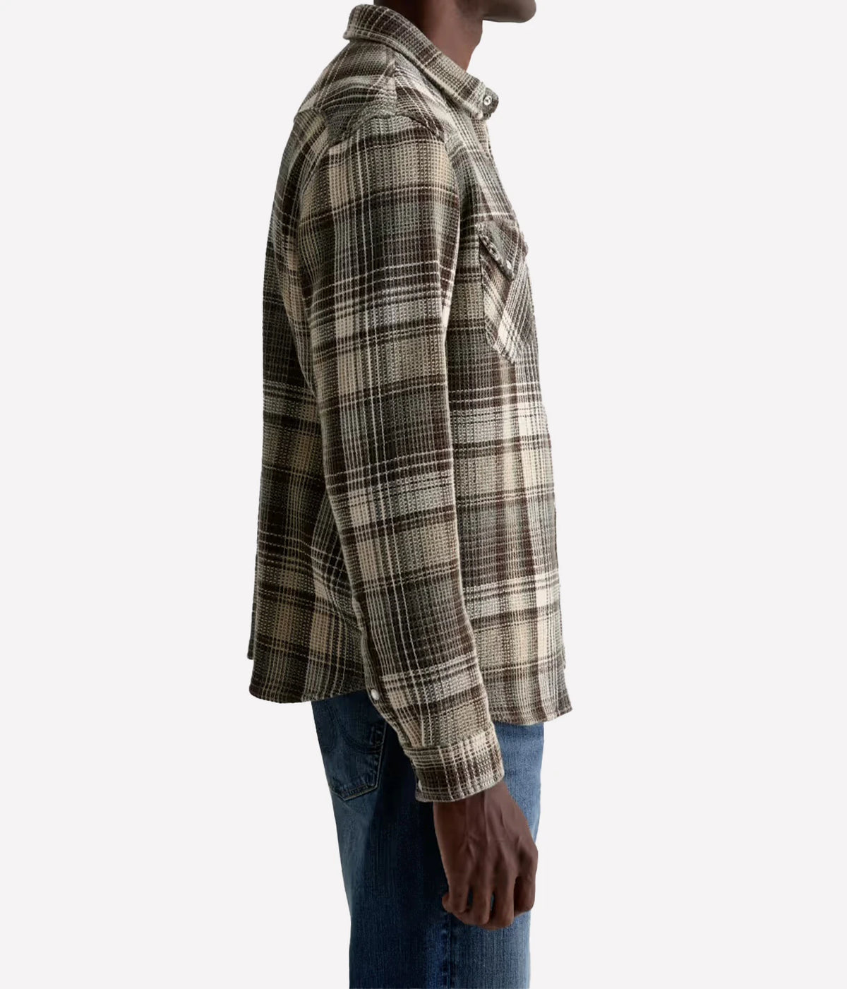 A man shown in side profile wearing a long-sleeve plaid flannel shirt in beige, brown, and black, paired with blue jeans, against a plain light gray background.