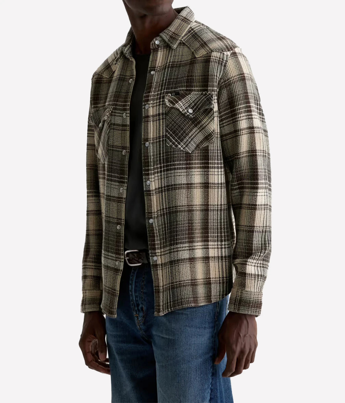 A man wearing a long-sleeve plaid button-up shirt in brown, beige, and black tones, layered over a black t-shirt and blue jeans, shown from the neck down on a light gray background.