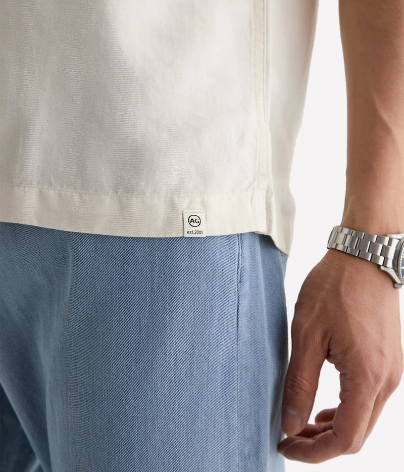 Close-up of a person wearing a light cream-colored short-sleeve shirt and light blue trousers, with a silver metal link watch on the wrist. The shirt features a small white brand tag near the hem that reads 'AQUA' with a circular logo.