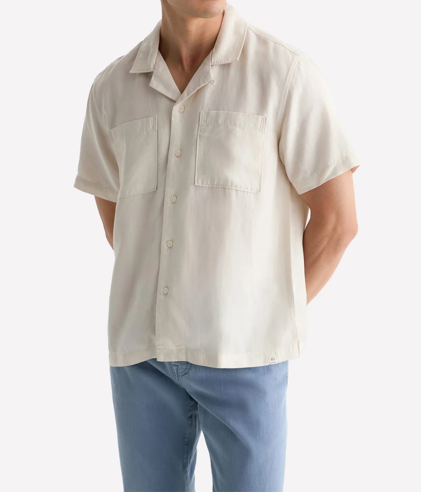 A man wearing a short-sleeve camp collar button-up shirt in off-white linen, paired with light blue jeans, shown from the neck down against a plain white background.