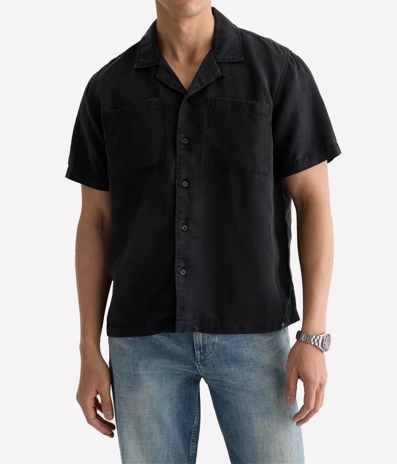 A man wearing a black short-sleeve camp collar button-up shirt and blue jeans, shown from the chin down against a light gray studio background.