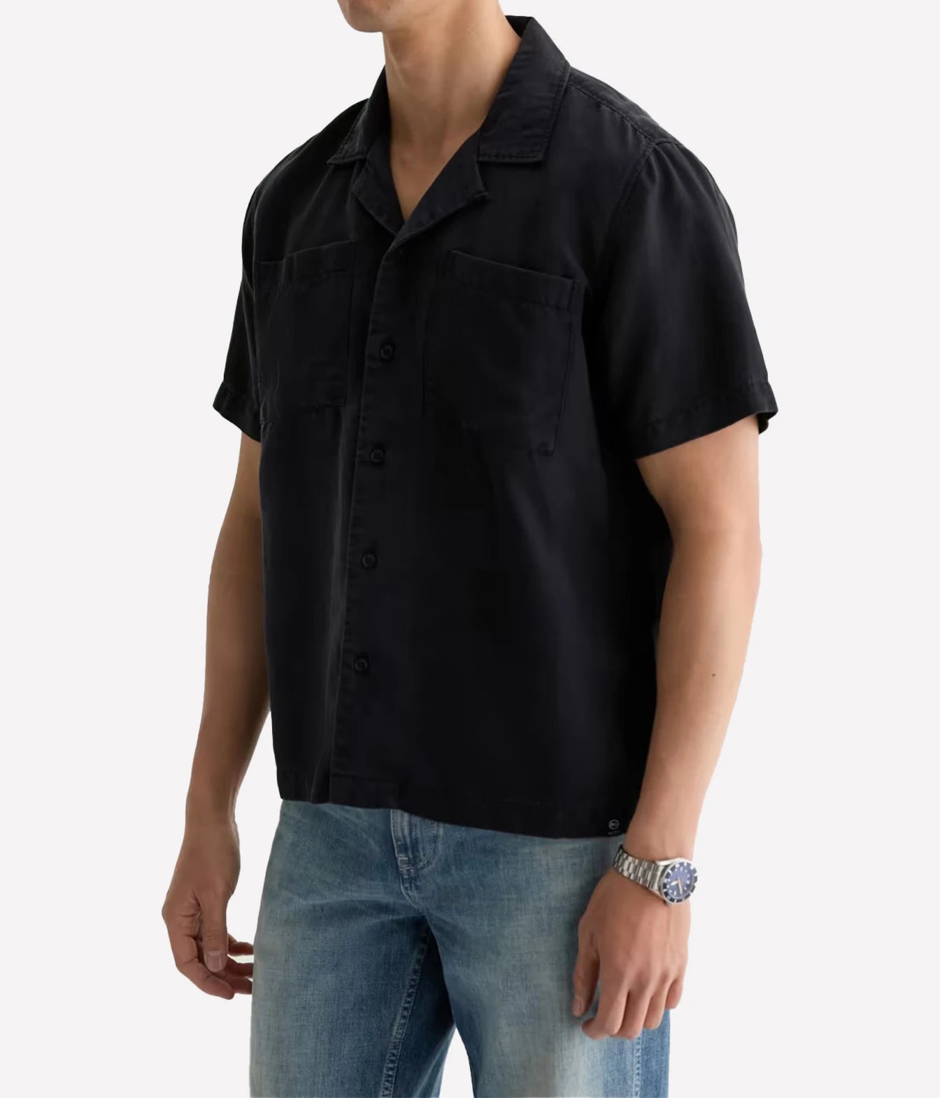 A man wearing a black short-sleeve button-up shirt with two chest pockets and blue jeans, shown from the neck down against a light gray background.