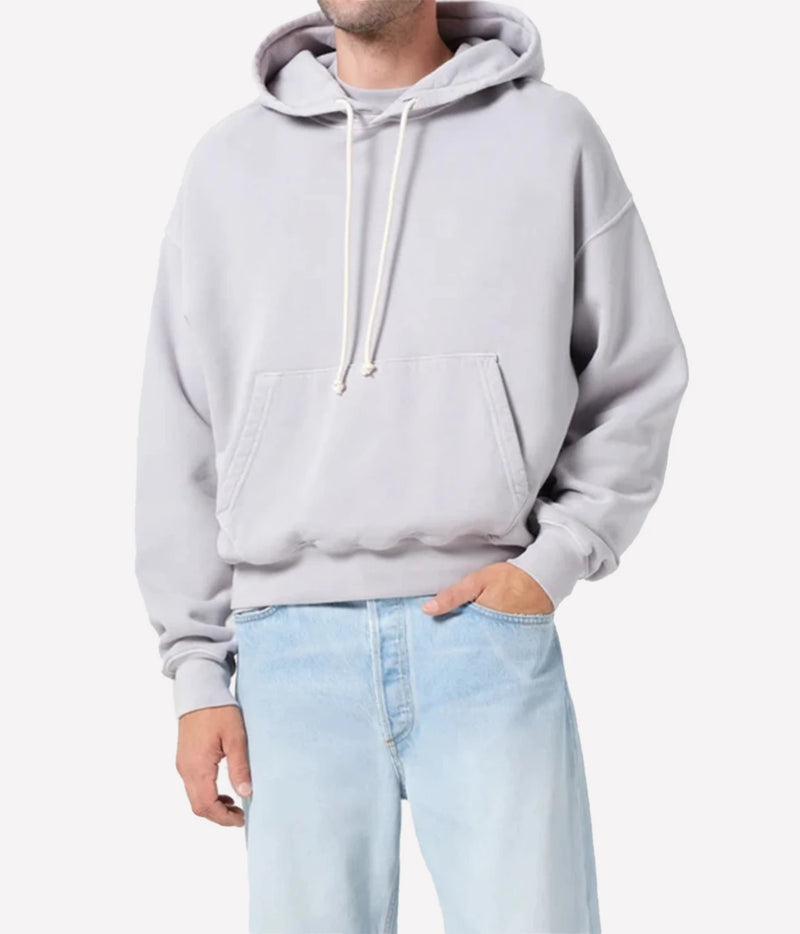A man wearing a light gray oversized pullover hoodie with white drawstrings and a front kangaroo pocket, paired with light blue jeans, against a plain light gray background.