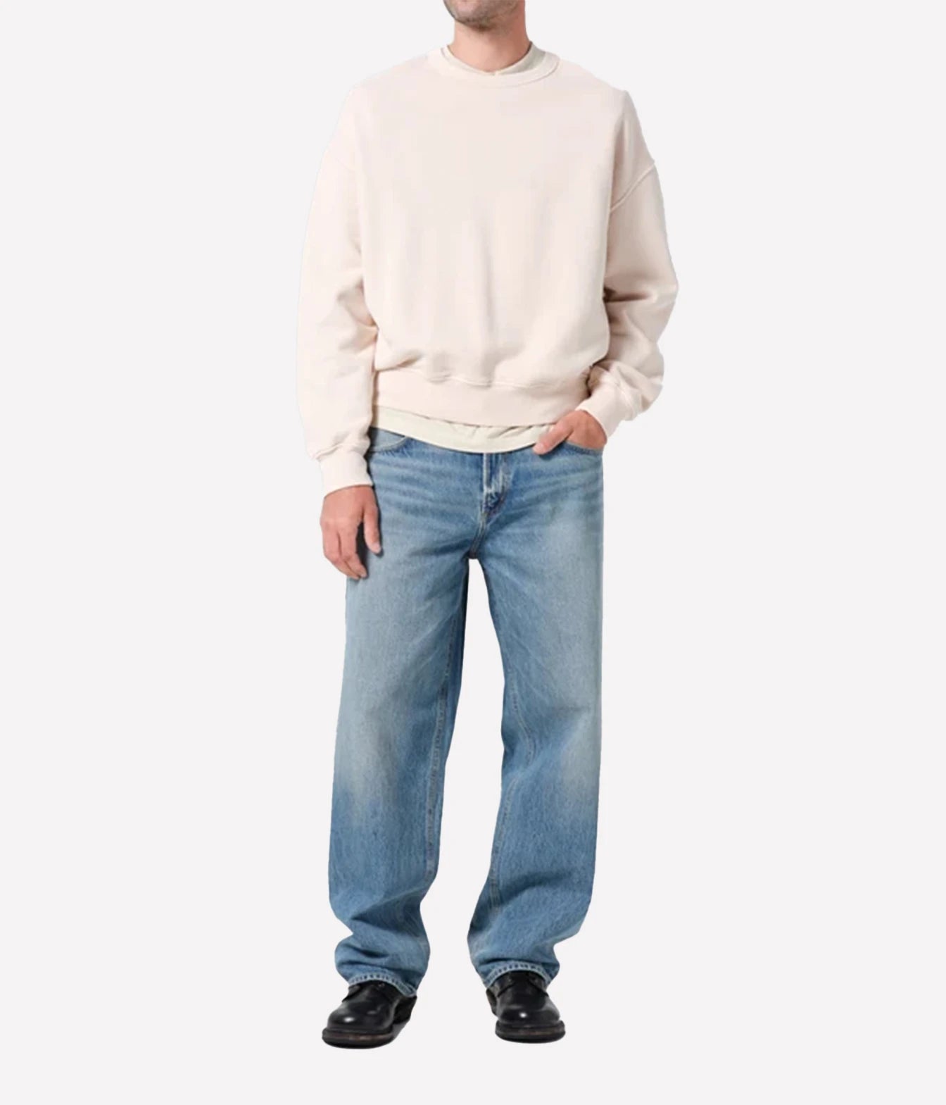 A model wearing a cream-colored crewneck sweatshirt, light-wash blue jeans, and black leather shoes, standing against a plain light gray background.