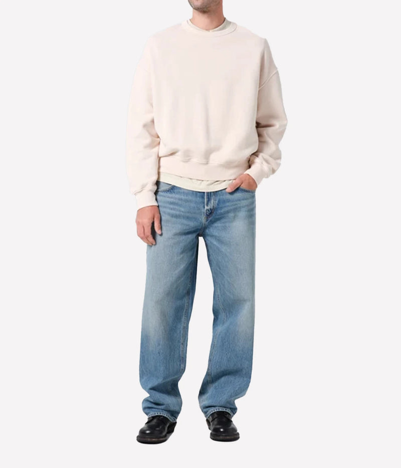 A model wearing a cream-colored crewneck sweatshirt, light-wash blue jeans, and black leather shoes, standing against a plain light gray background.