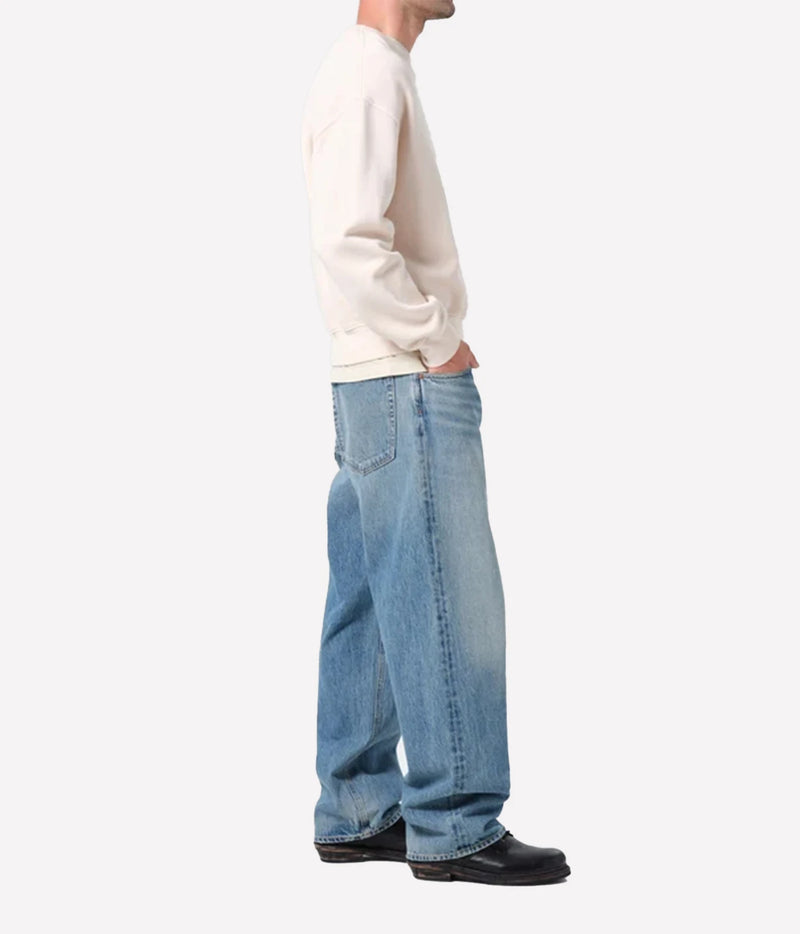 A side profile view of a person wearing light-wash blue wide-leg jeans, a cream sweatshirt, and black shoes against a plain light gray background.