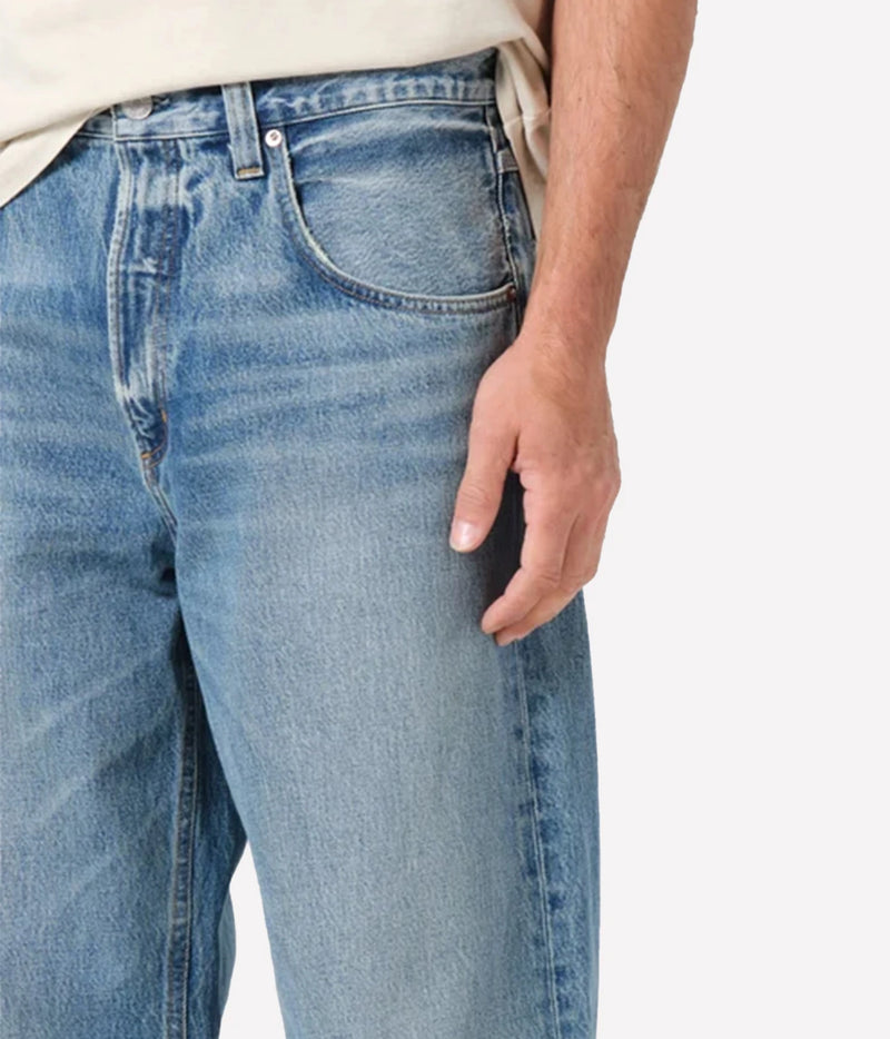 Close-up of a person wearing light blue denim jeans with a beige shirt partially visible, against a plain light background.