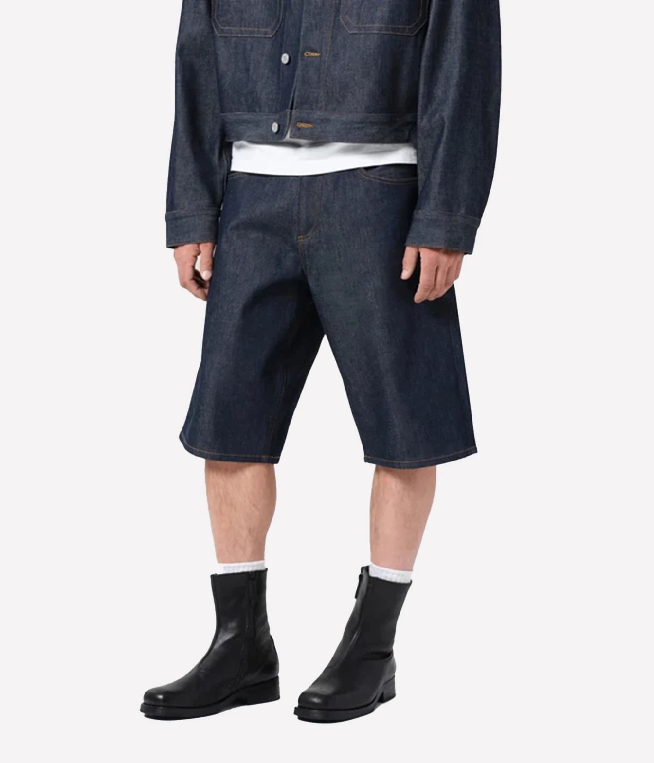 A model wearing dark indigo denim shorts, a matching dark denim jacket, a white t-shirt, white socks, and black leather ankle boots, shown from the waist down on a light gray background.
