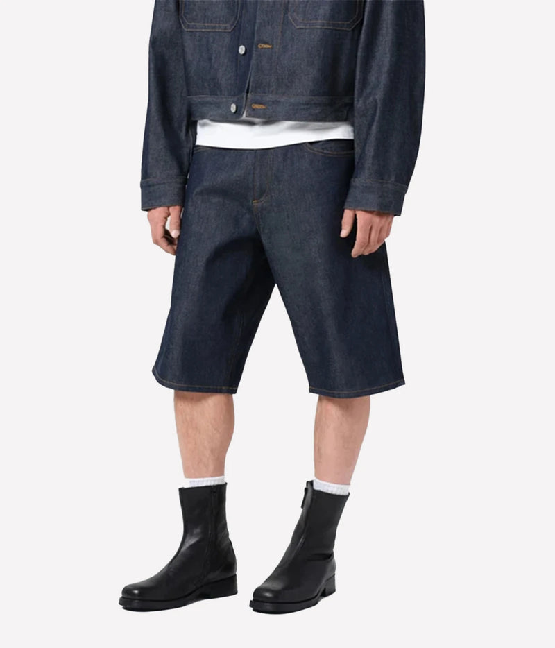 A model wearing dark indigo denim shorts, a matching dark denim jacket, a white t-shirt, white socks, and black leather ankle boots, shown from the waist down on a light gray background.