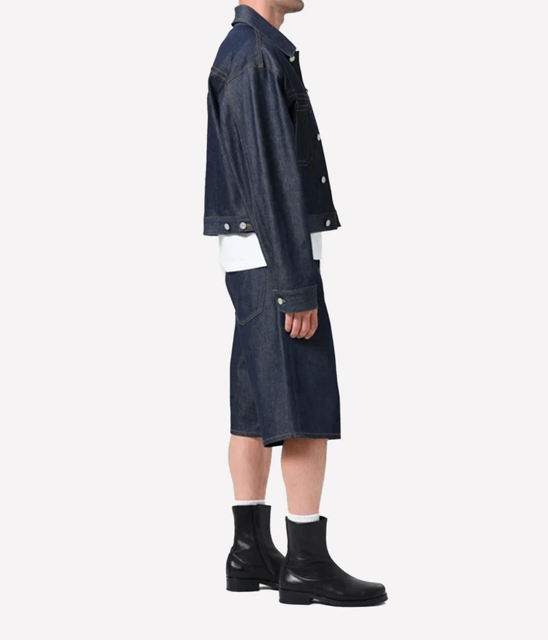 A model shown from the side wearing a dark indigo denim jacket, matching denim shorts, a white shirt, and black leather ankle boots against a plain light gray background.