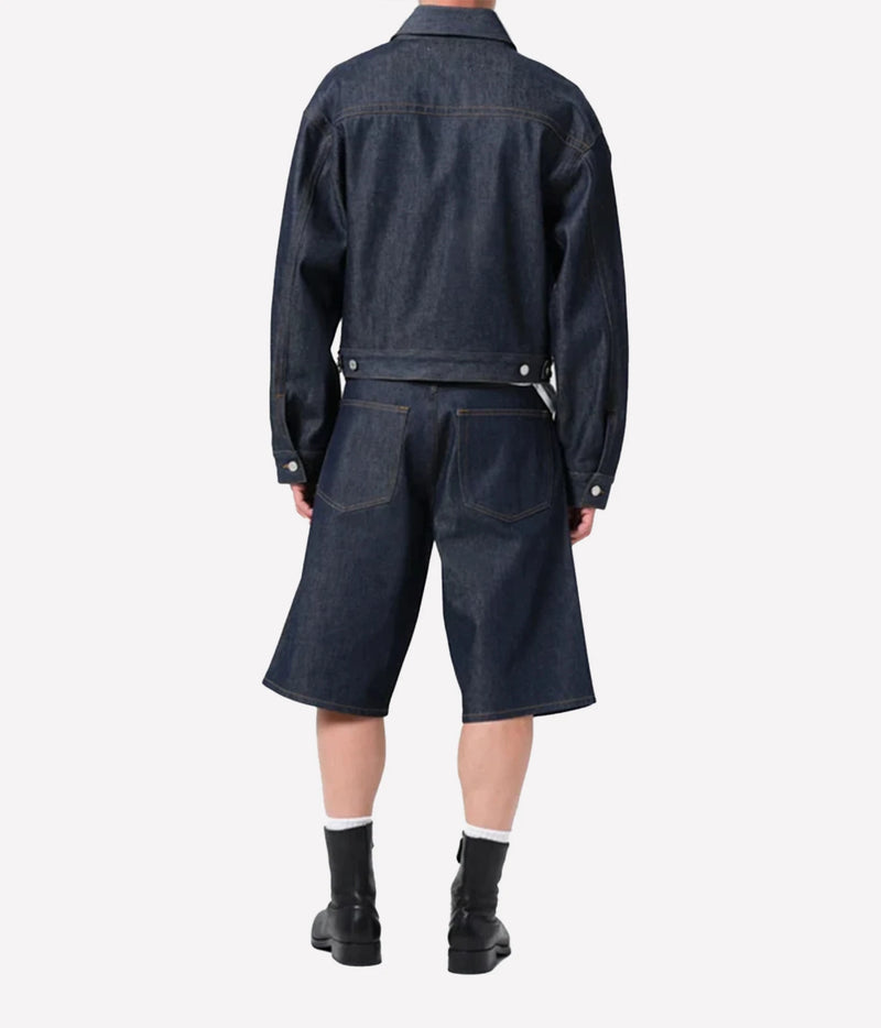 A model shown from the back wearing a dark indigo denim jacket and matching cropped denim shorts, paired with black boots and white socks, against a plain light gray background.