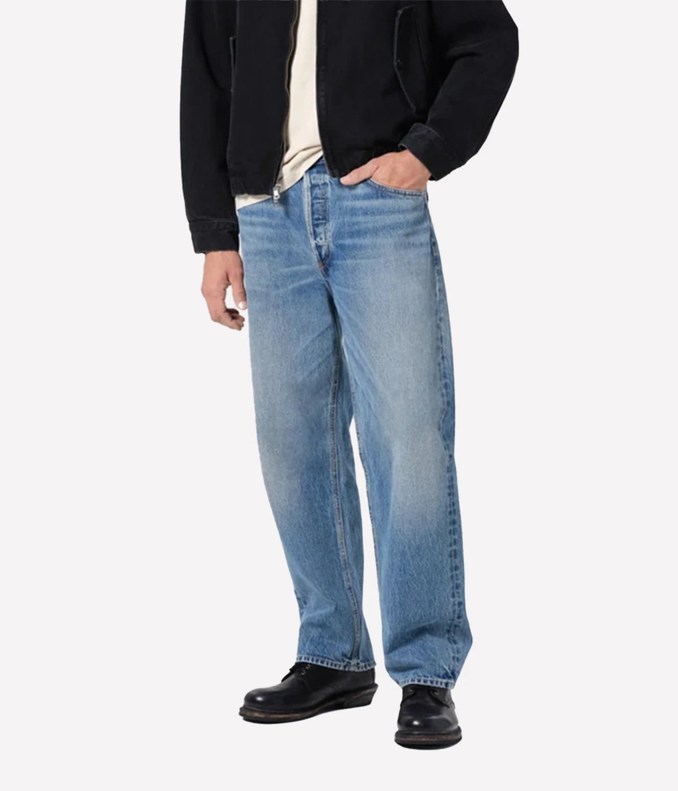 A person wearing light blue relaxed-fit denim jeans, a black jacket, a cream shirt, and black leather shoes, standing against a light gray studio background.