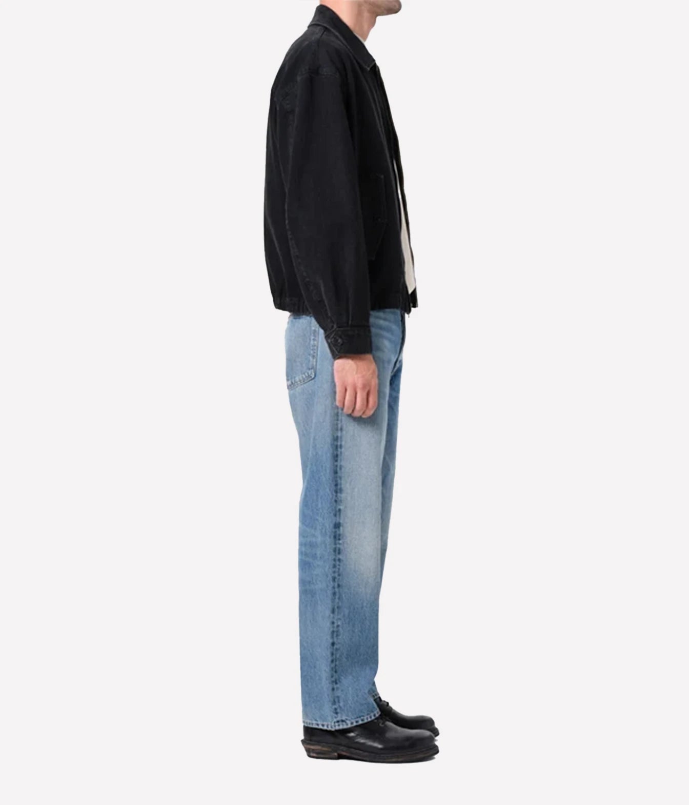 A side profile view of a person wearing a black jacket, light blue jeans, and black leather shoes against a plain white background.