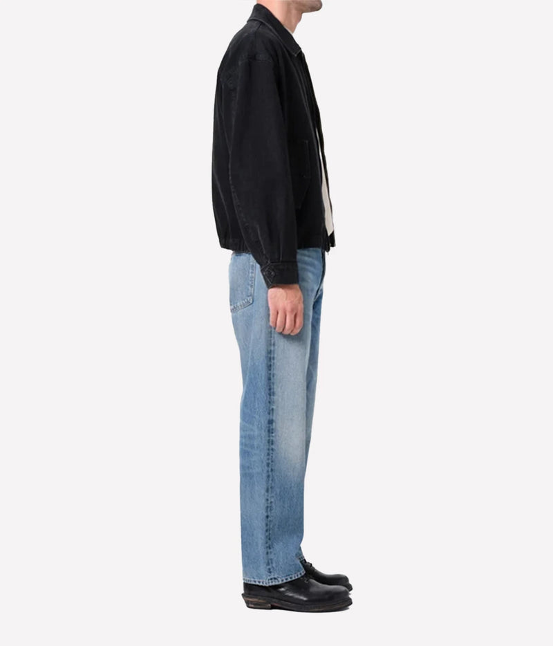A side profile view of a person wearing a black jacket, light blue jeans, and black leather shoes against a plain white background.