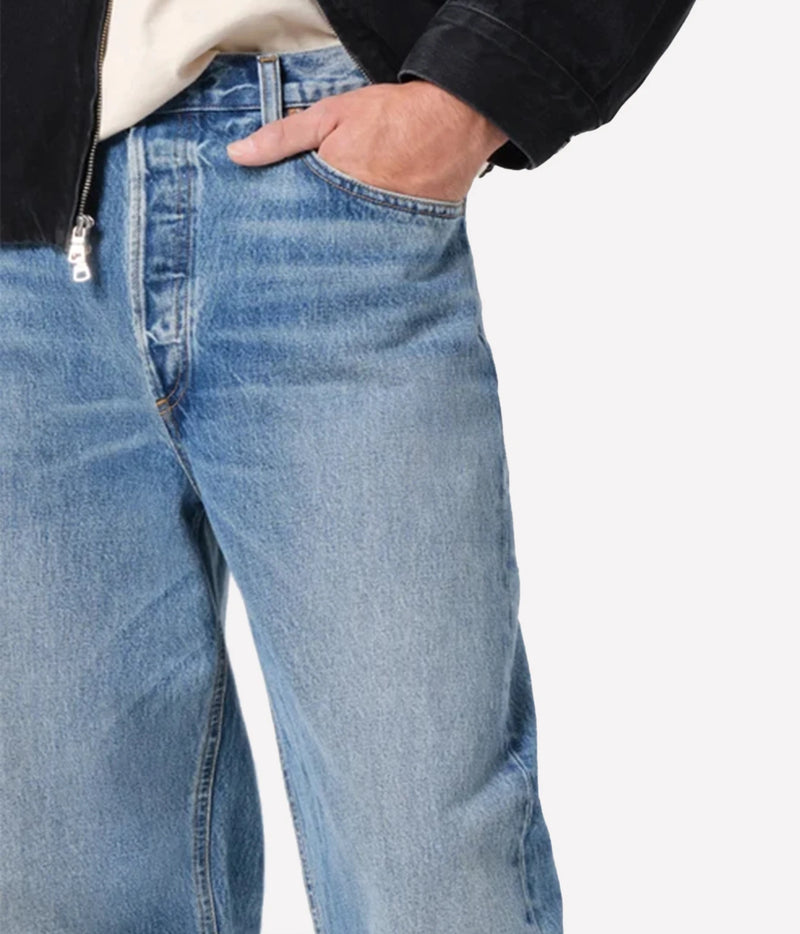 Close-up of a person wearing light blue denim jeans with a hand in the front pocket, styled with a cream shirt and a black jacket against a light gray background.