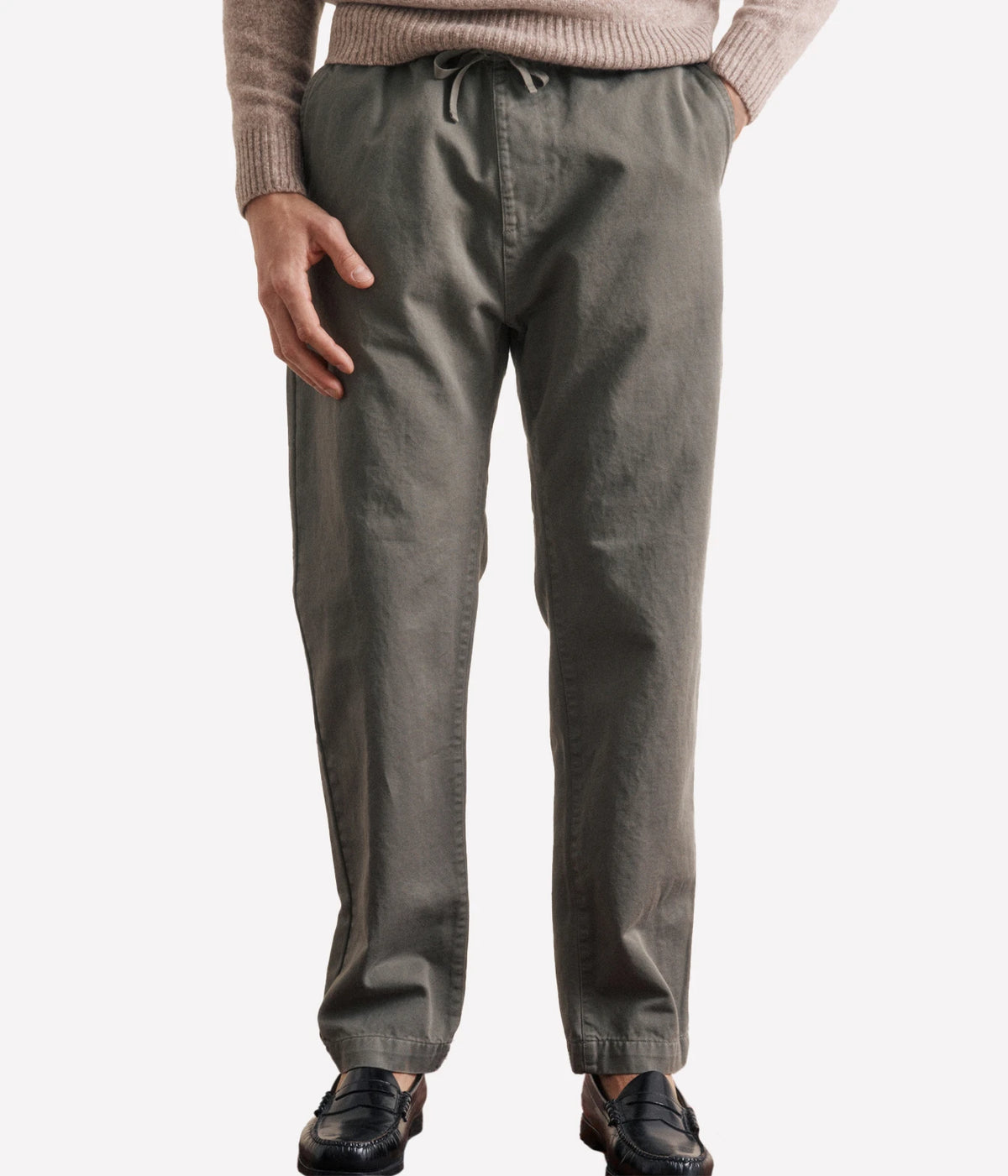 A person wearing dark olive green drawstring trousers, a beige ribbed sweater, and black loafers, shown from the waist down against a light grey studio background.