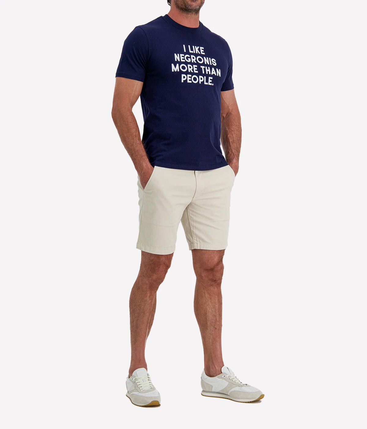 Man wearing The Refined Spirit I Like Negronis More Than People Tee in Navy with stone shorts and white sneakers, full-length front view with hand in pocket
