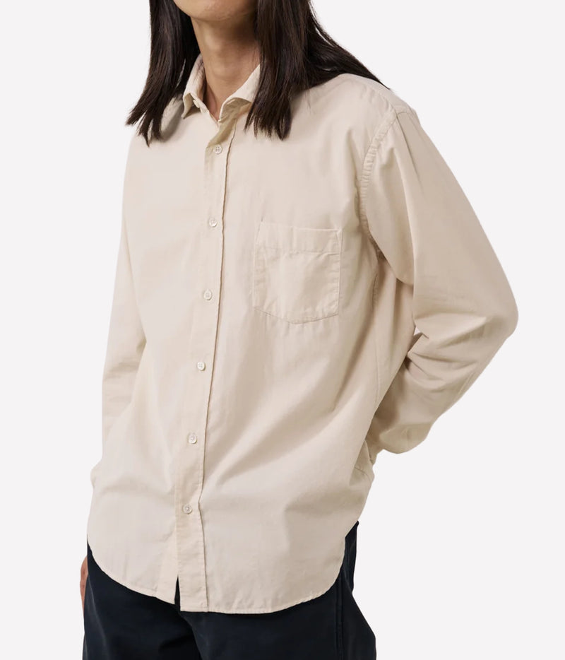 A person wearing a light beige long-sleeve button-up shirt with a chest pocket, paired with dark pants, against a plain light grey background.