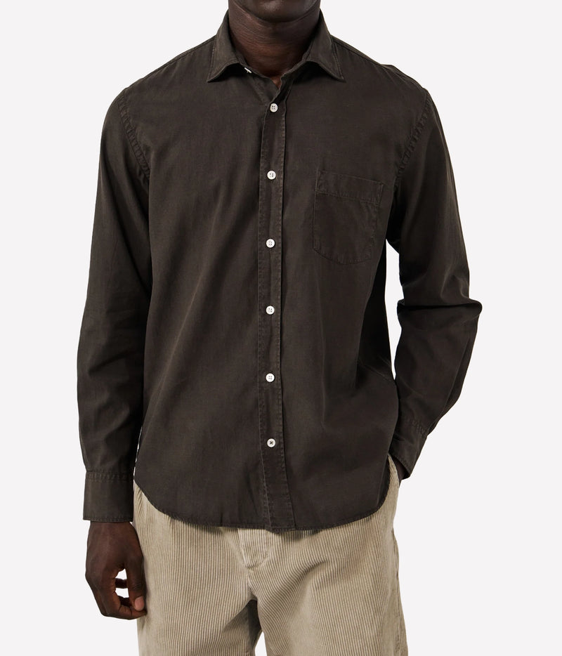 A man wearing a dark brown long-sleeve button-down shirt with a chest pocket, paired with beige corduroy trousers, against a plain light grey background.