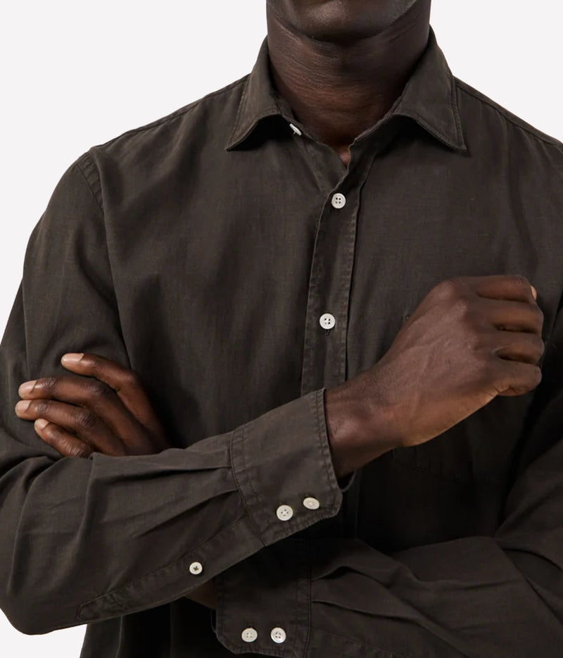 A close-up of a man wearing a dark brown long-sleeve button-down shirt, showing the collar, front buttons, and crossed arms against a light grey background.