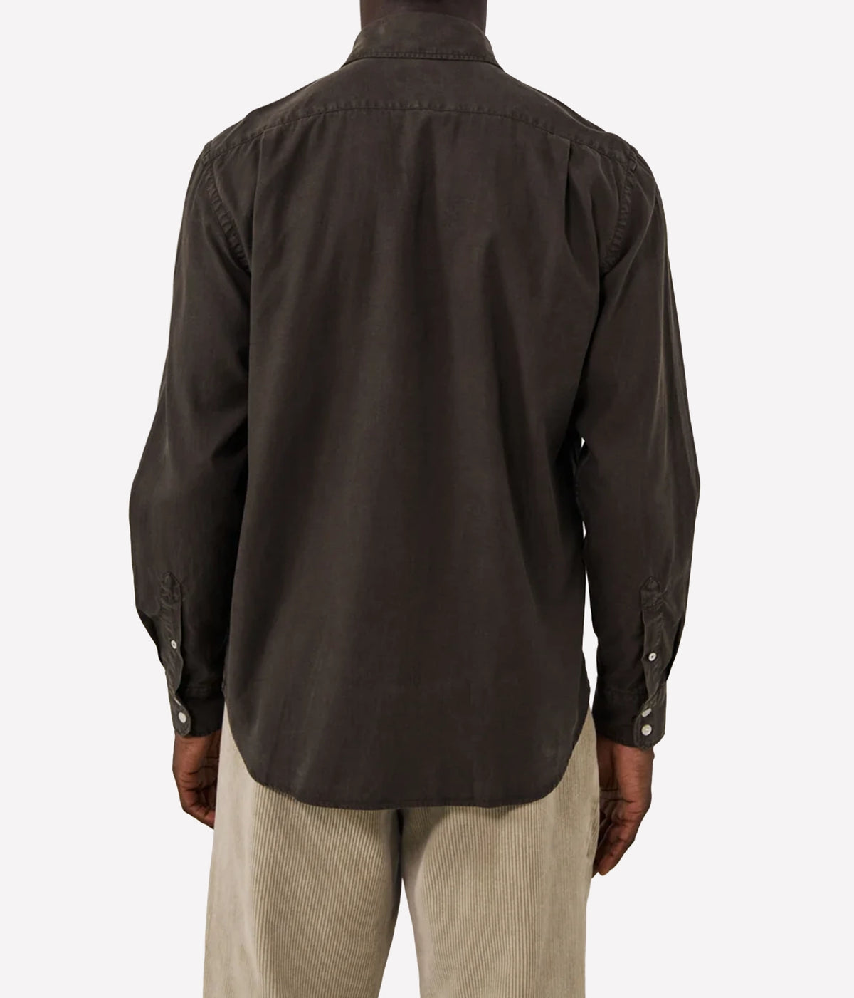 A man viewed from the back wearing a dark brown long-sleeve button-down shirt and light beige corduroy trousers, against a plain light grey background.