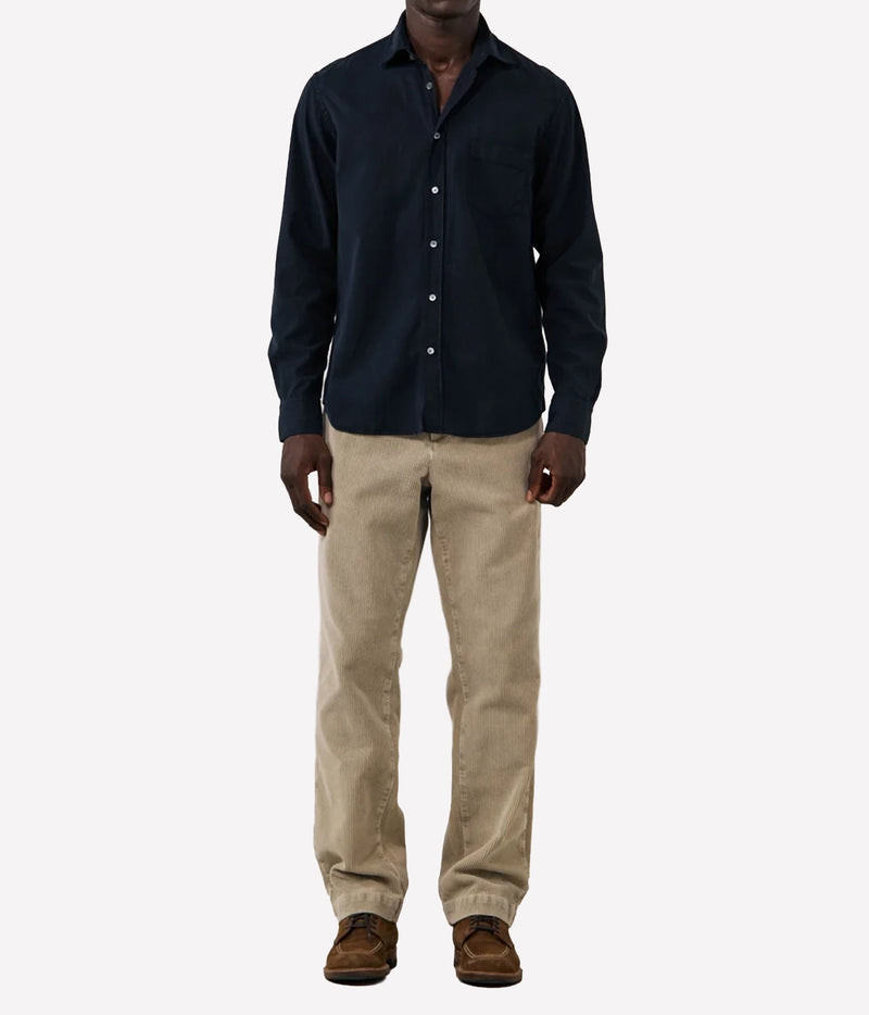 A man wearing a dark navy blue long-sleeve button-down shirt, beige trousers, and brown shoes, standing against a plain light grey background.