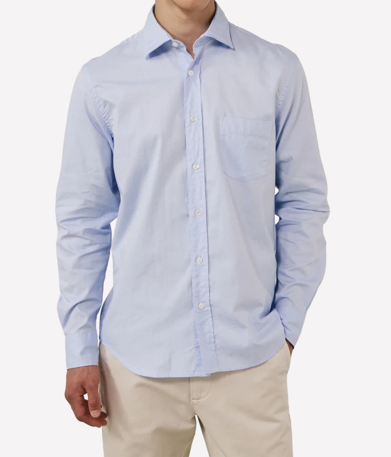 A man wearing a light blue long-sleeve button-down shirt with a chest pocket, paired with beige trousers, shown against a plain light grey background.