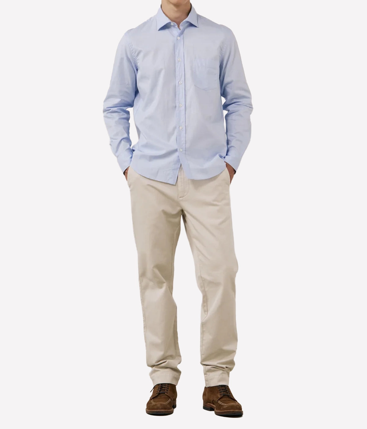 A man wearing a light blue long-sleeve button-down shirt, beige trousers, and brown shoes, standing against a plain light grey background.