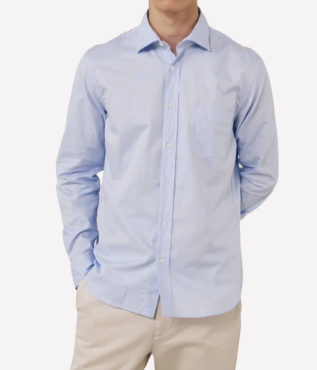 A man wearing a light blue long-sleeve button-down shirt with a chest pocket, paired with beige trousers, shown against a plain light grey background.