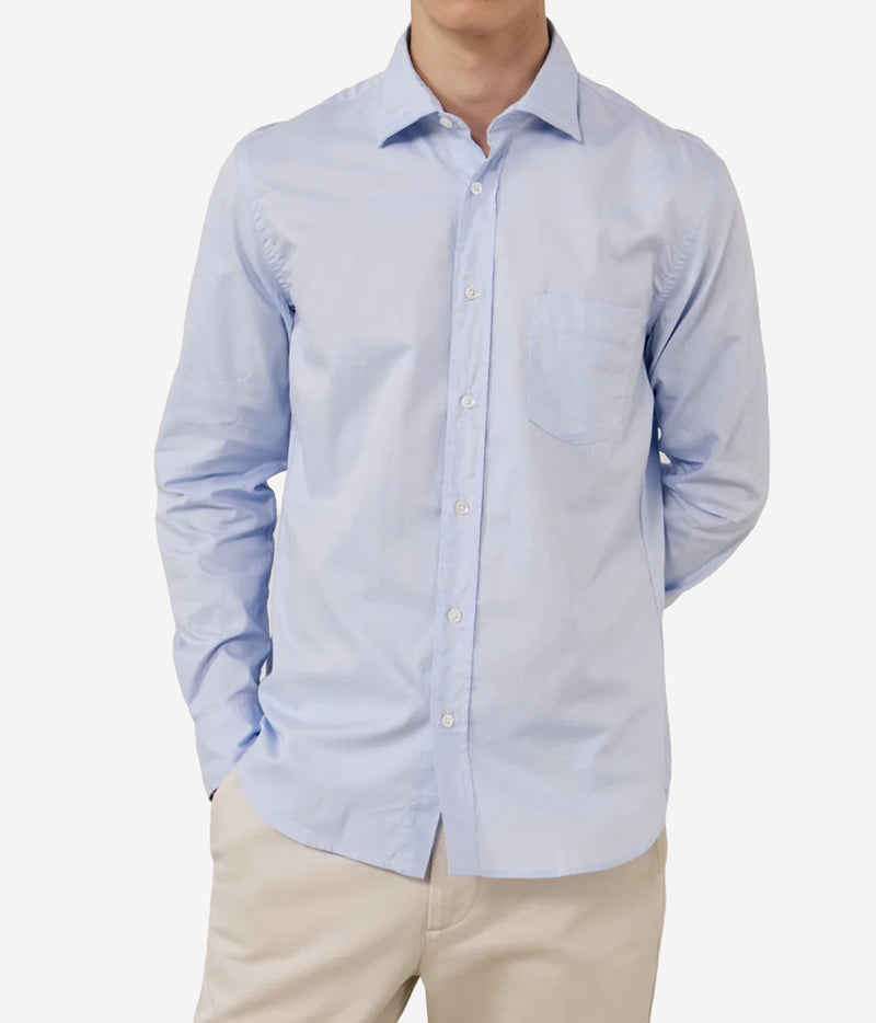 A man wearing a light blue long-sleeve button-down shirt with a chest pocket, paired with beige trousers, shown against a plain light grey background.