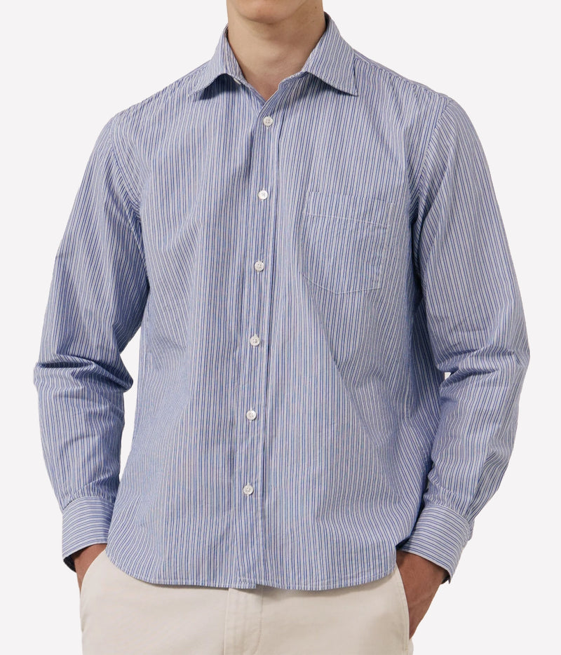 A man wearing a blue and white vertically striped long-sleeve button-down shirt with a chest pocket, paired with light beige trousers.