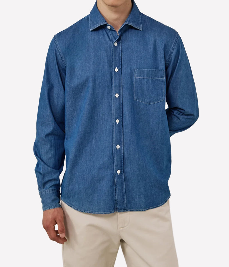 A man wearing a blue long-sleeve denim button-up shirt with a chest pocket, paired with beige pants, shown against a plain light grey background.