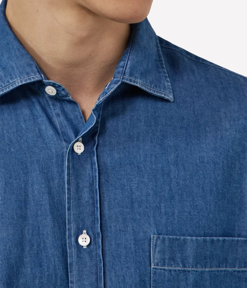 Close-up of a person wearing a blue denim or chambray button-up shirt with white buttons and a chest pocket, against a light neutral background.