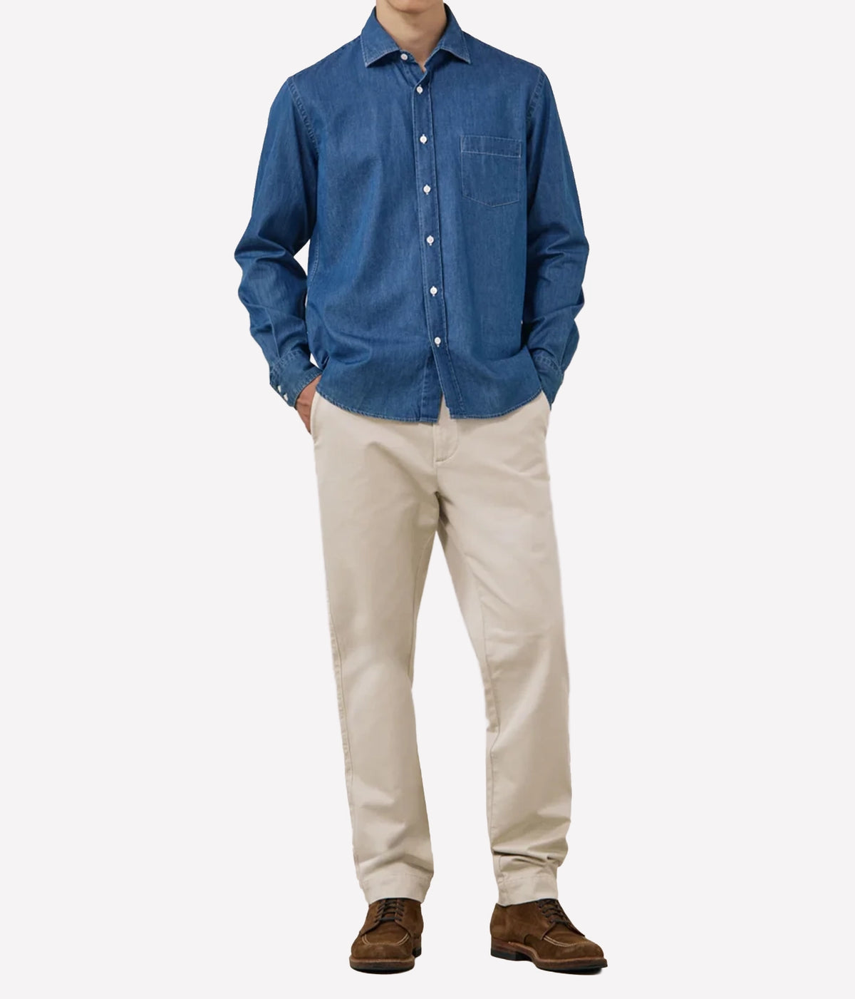 A man wearing a blue long-sleeve button-up shirt, beige trousers, and brown shoes, standing against a light grey studio background.