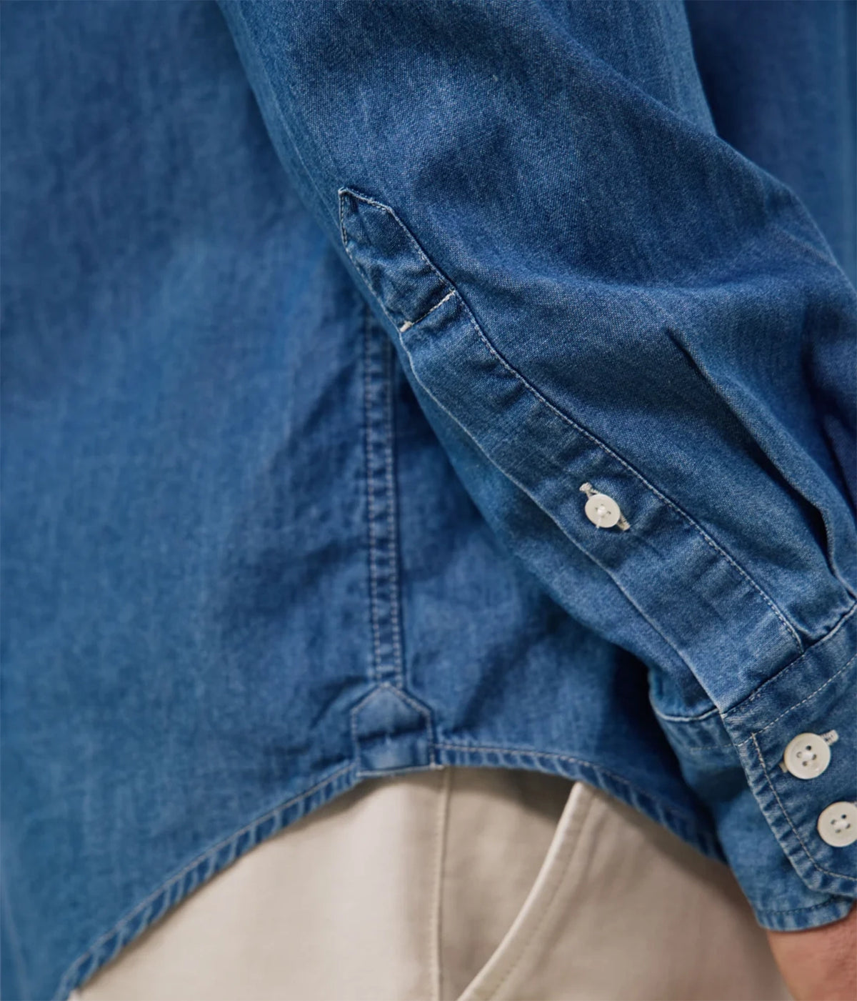 Close-up of a blue denim shirt with a buttoned cuff and light beige pants visible underneath.