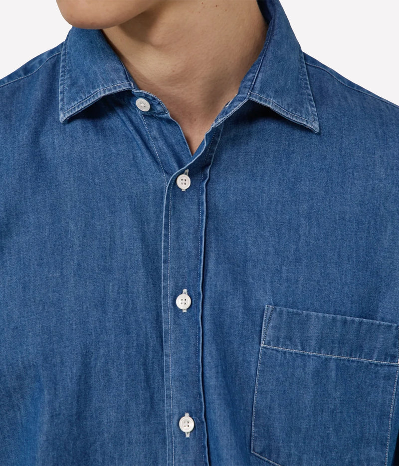 Close-up of a person wearing a blue denim or chambray button-up shirt with a collar, white buttons, and a chest pocket against a light grey background.