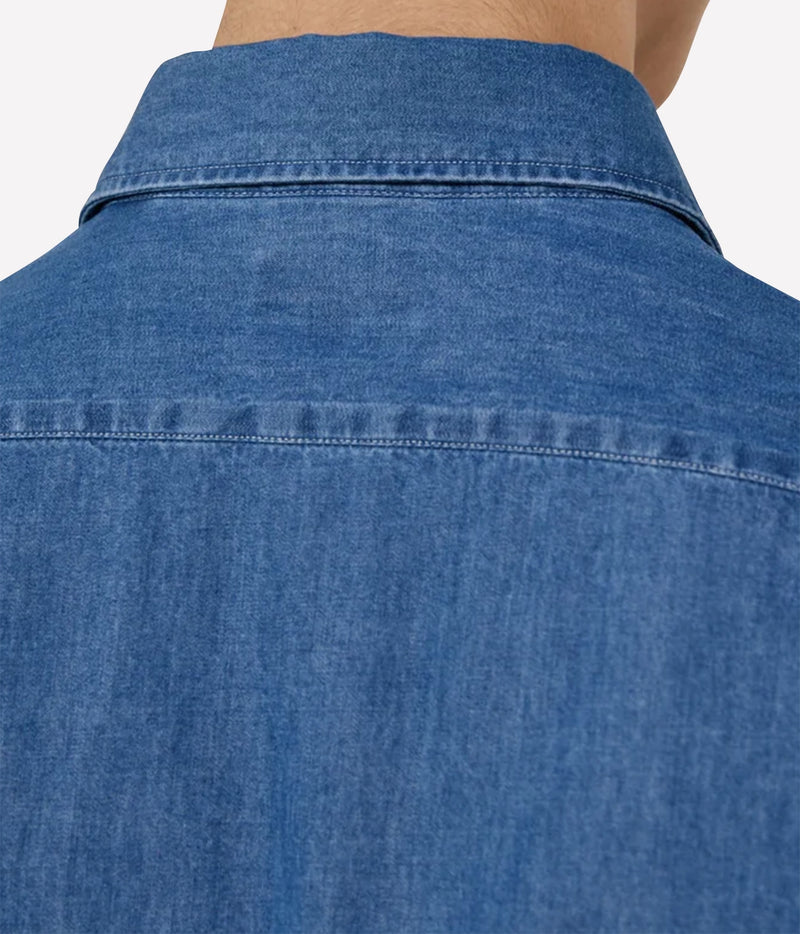 Close-up of the back of a blue denim shirt worn by a model, showing the collar, shoulder seam, and fabric texture against a light neutral background.